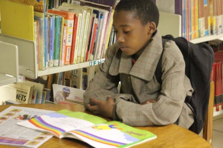 Jeune élève assis à une table de bibliothèque, lisant un livre illustré, rangées d’ouvrages en arrière-plan.