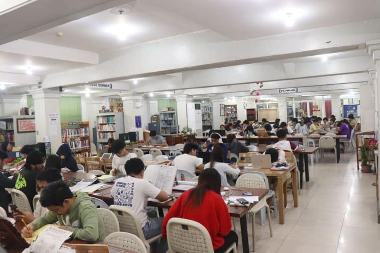 Grande salle de bibliothèque remplie d’étudiants concentrés à des tables, cahiers ouverts et ordinateurs posés.