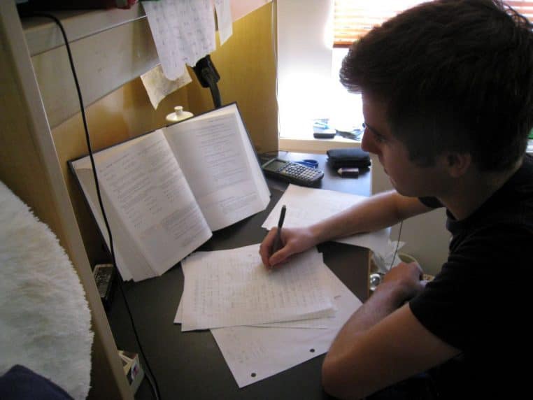 Étudiant penché sur ses feuilles de cours à son bureau, stylo en main, préparant des examens de fin d’année.