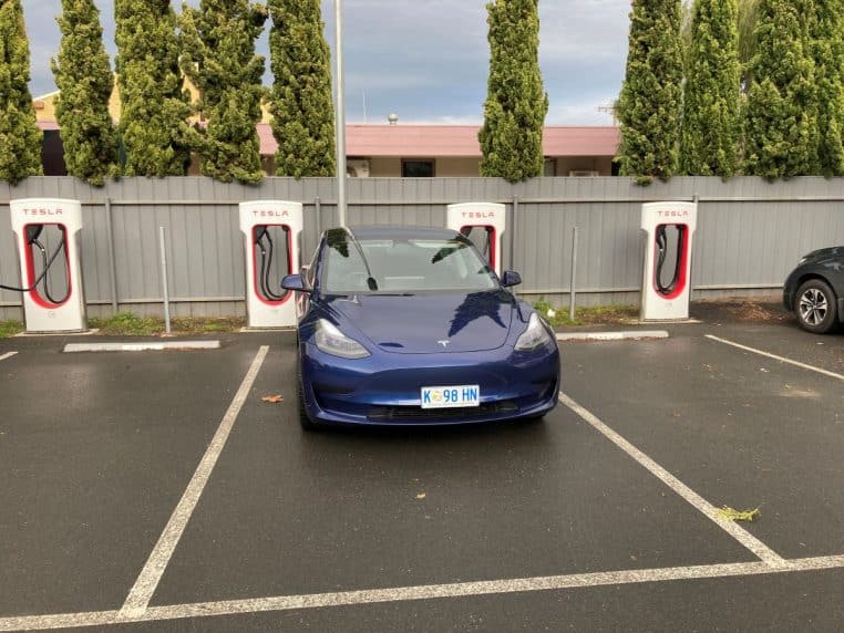 Tesla Model 3 stationnée à un Superchargeur, bornes visibles, lumière du matin, décor routier