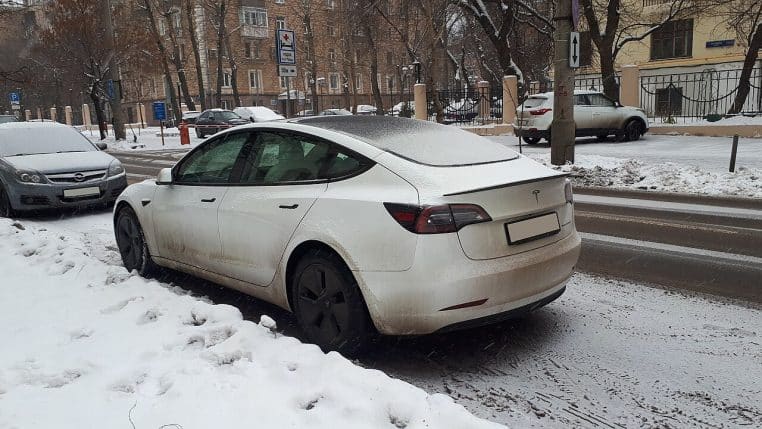 Tesla Model S blanche stationnée au bord d’une rue enneigée, vue arrière, hiver, traces de neige et route mouillée.