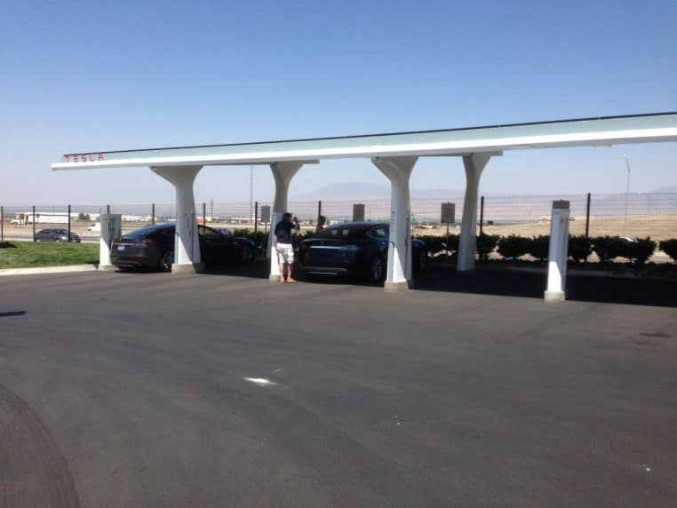 Station de recharge Tesla avec ombrière solaire, plusieurs voitures alignées, ciel bleu dégagé