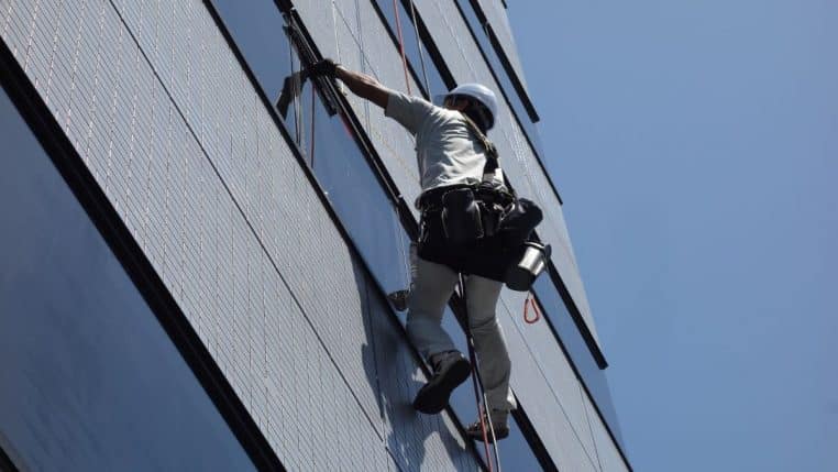 Agent en train de nettoyer les vitres d’un immeuble moderne, suspendu avec harnais, vue en contre-plongée.