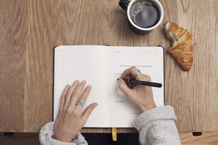Vue du dessus d’un planner ouvert, une main écrit au stylo, tasse de café et croissant sur la table