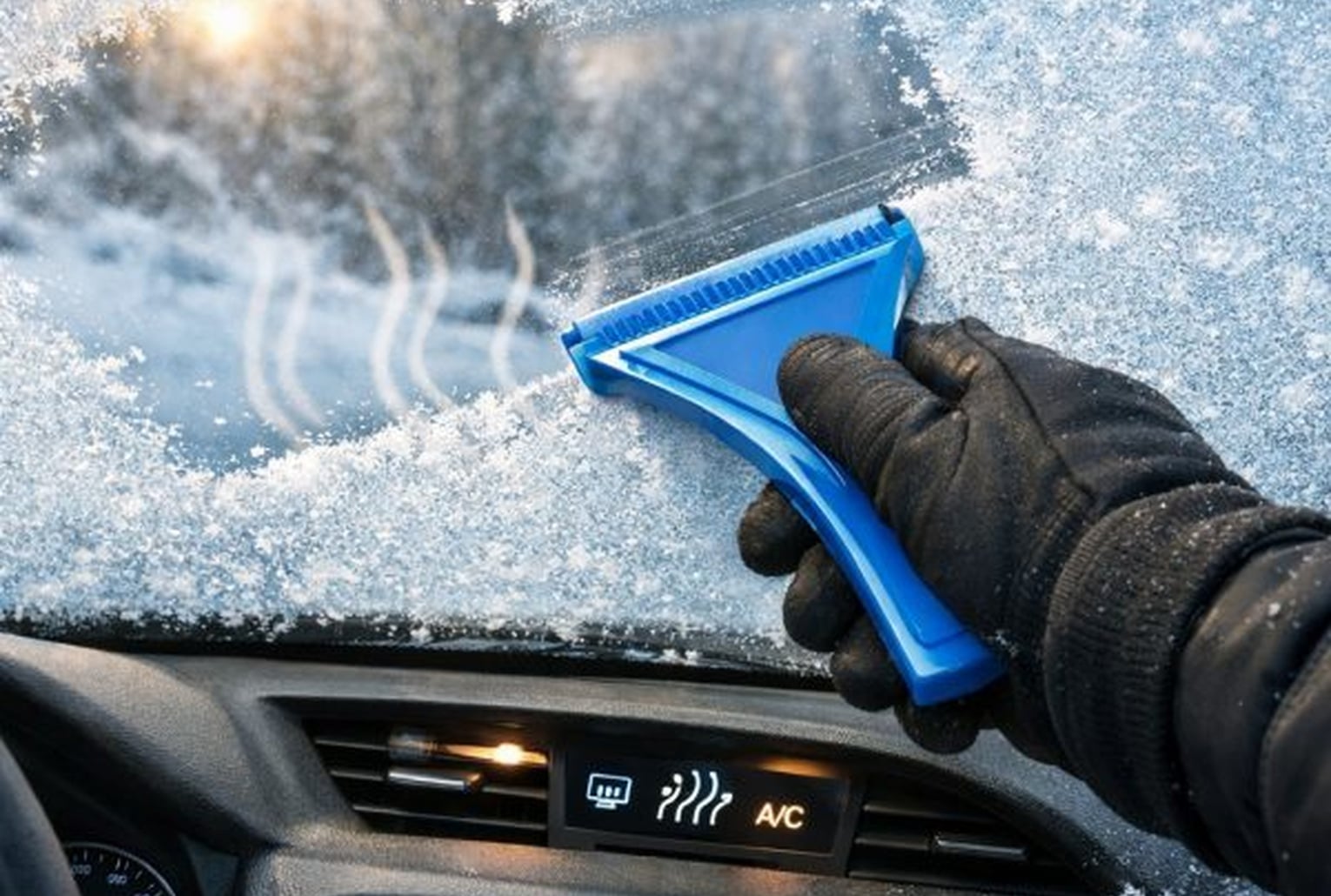 Main gantée grattant un pare-brise givré avec une raclette en plastique, chauffage/désembuage activé à l’intérieur.