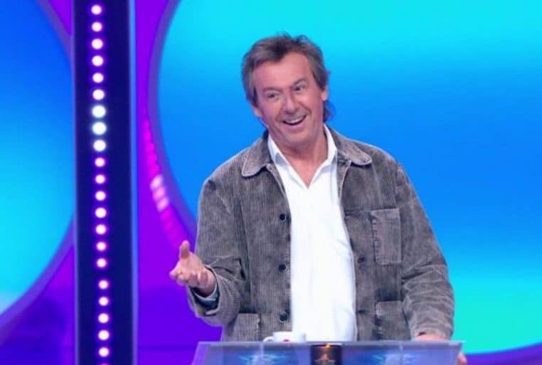 Jean-Luc Reichmann souriant sur le plateau des « 12 Coups de midi », devant le pupitre, en plan large.