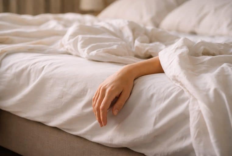 Main d’une personne dépassant du bord d’un lit défait, draps blancs froissés, lumière douce du matin.