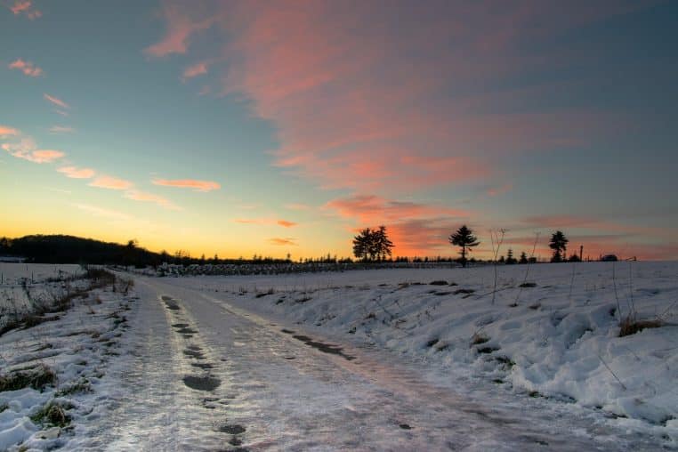 Route verglacée en campagne au coucher du soleil, bande brillante de glace et champs enneigés autour.
