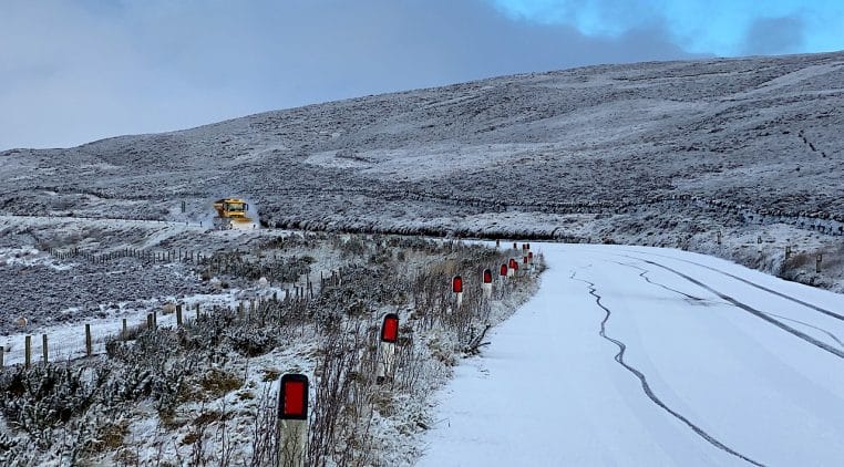 Route de montagne enneigée et glacée avec balises rouges, virage serré et déneigeuse au loin.