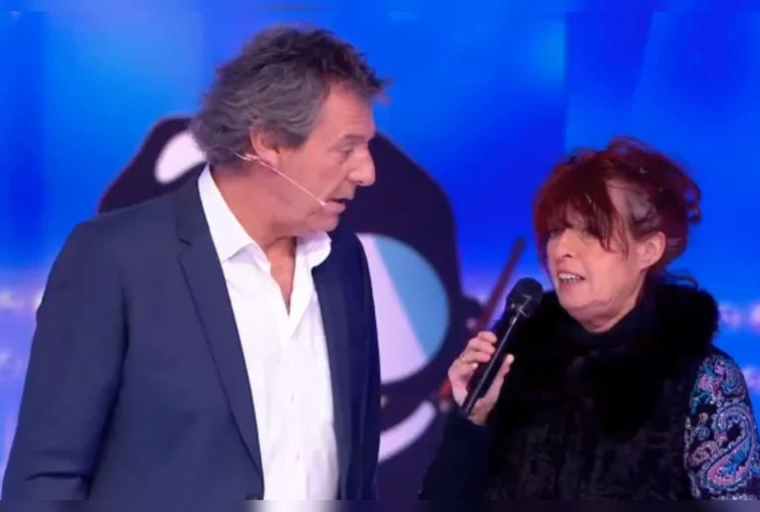 Jean-Luc Reichmann sur le plateau des 12 Coups de midi aux côtés de Zette tenant un micro, en arrière-plan bleu.