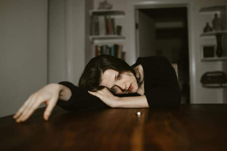 A thoughtful woman rests her head on a table in a dimly lit room, conveying a reflective mood.
