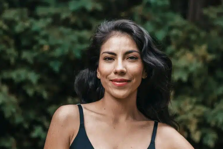 A close-up portrait of a smiling woman with black hair outdoors against lush greenery.