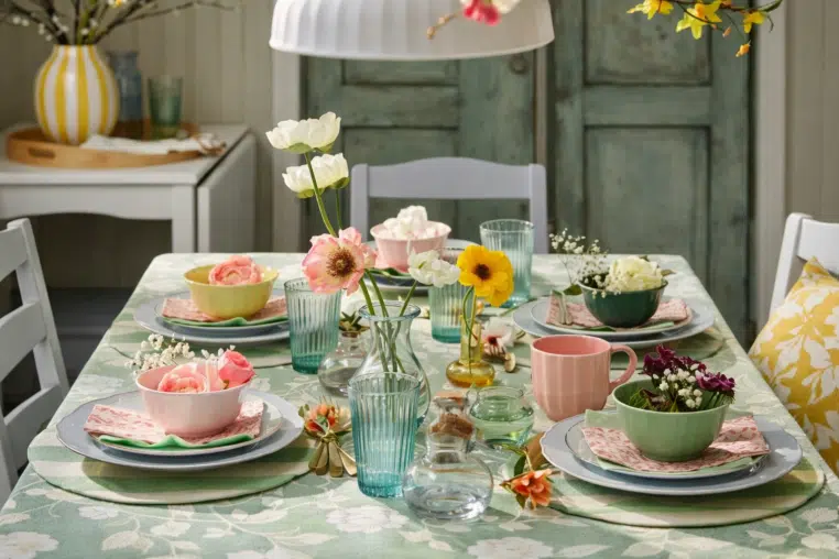 Table printanière pastel avec bols colorés, verres striés et fleurs sur nappe végétale, ambiance IKEA.