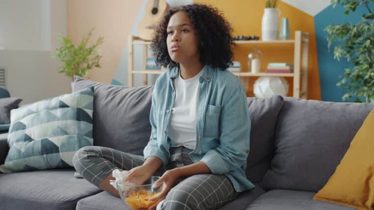 A young woman sits on a couch eating chips.