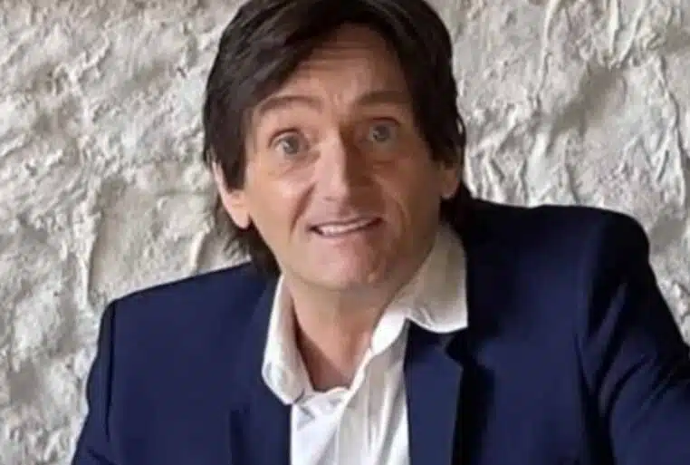 Pierre Palmade assis, costume bleu et chemise blanche, devant un mur en pierre claire.