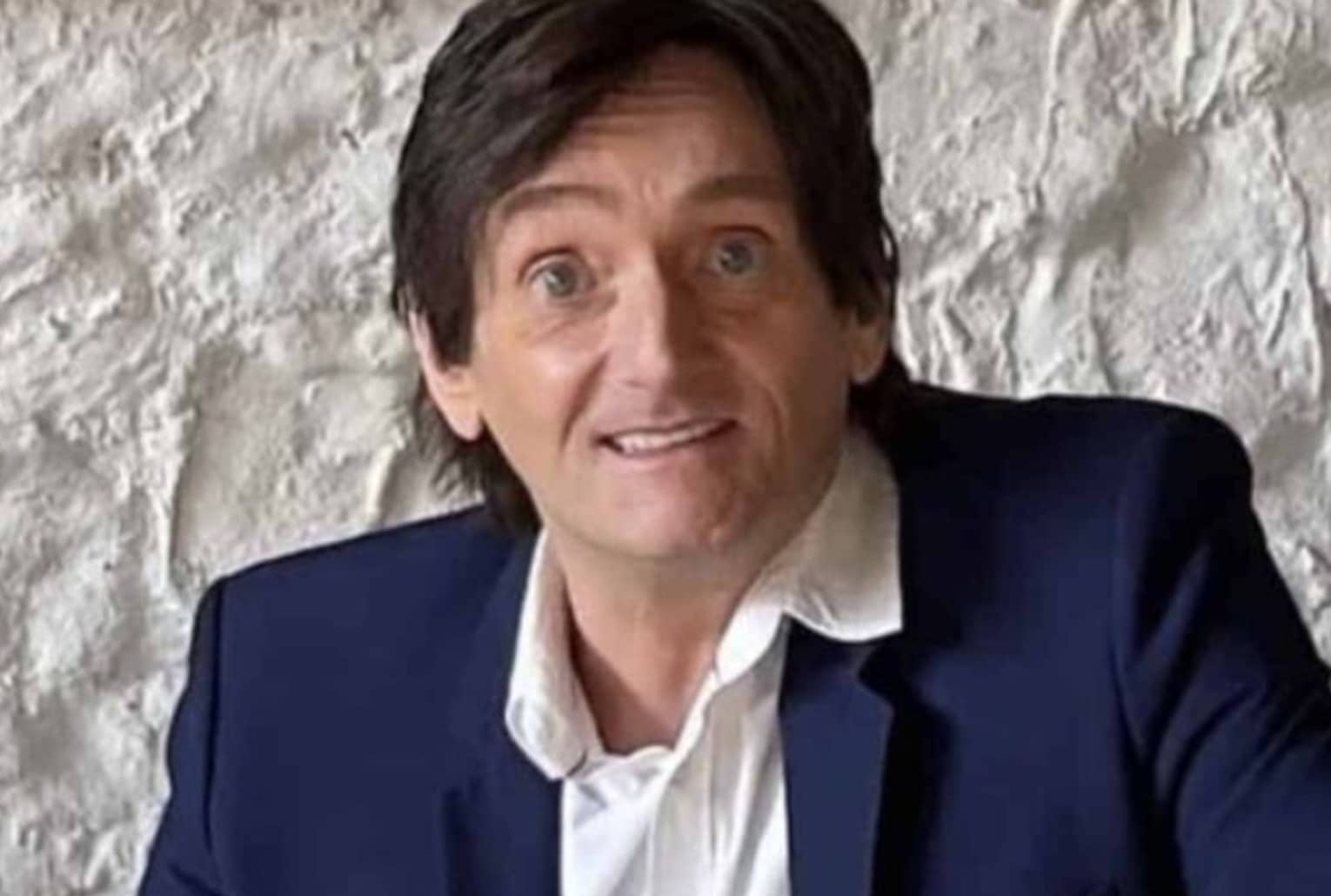 Pierre Palmade assis, costume bleu et chemise blanche, devant un mur en pierre claire.