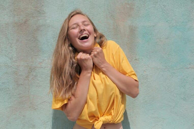 A cheerful woman in a yellow top laughing joyfully against a pastel-colored wall.