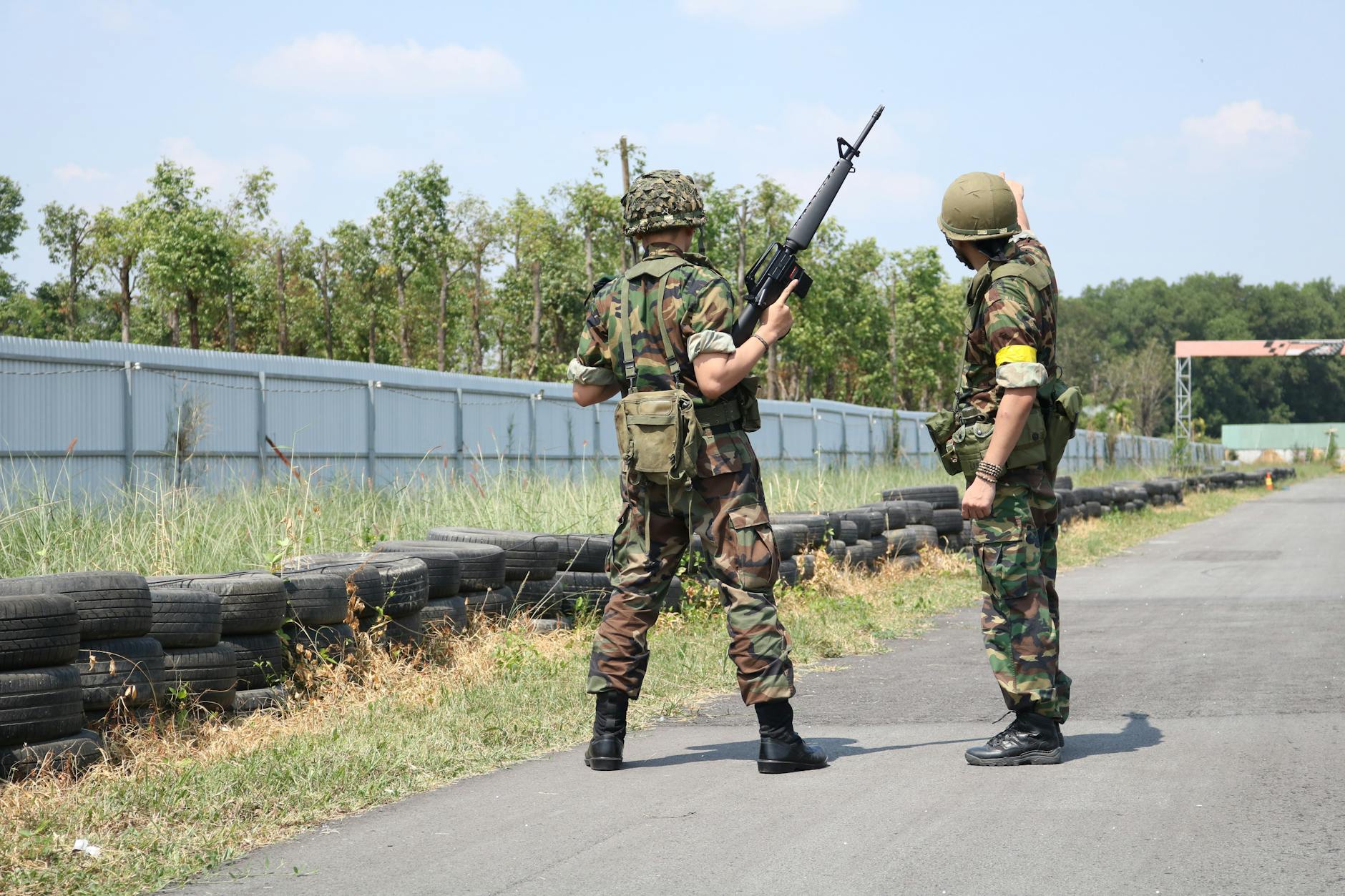 Two soldiers in camouflage uniforms with weapons stand outside on a training field.