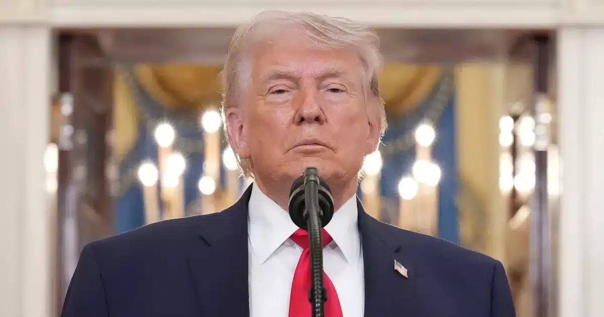 Donald Trump en costume sombre devant un micro, expression sérieuse, Maison Blanche en arrière-plan
