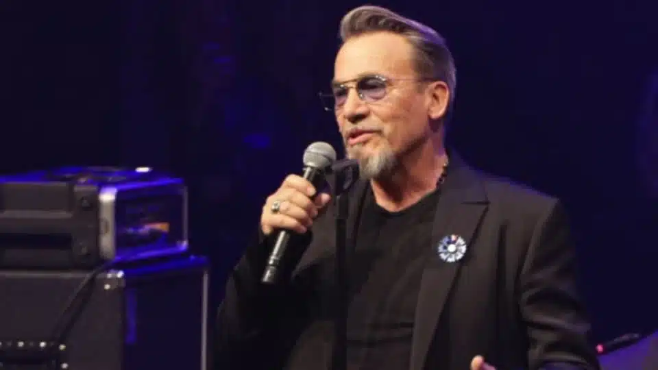 Florent Pagny tenant un microphone sur scène lors d'un concert, habillé en noir avec ses lunettes teintées