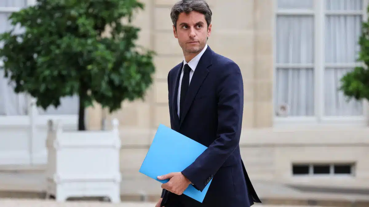 Gabriel Attal en costume sombre tenant un dossier bleu dans la cour de l'Élysée, regard déterminé