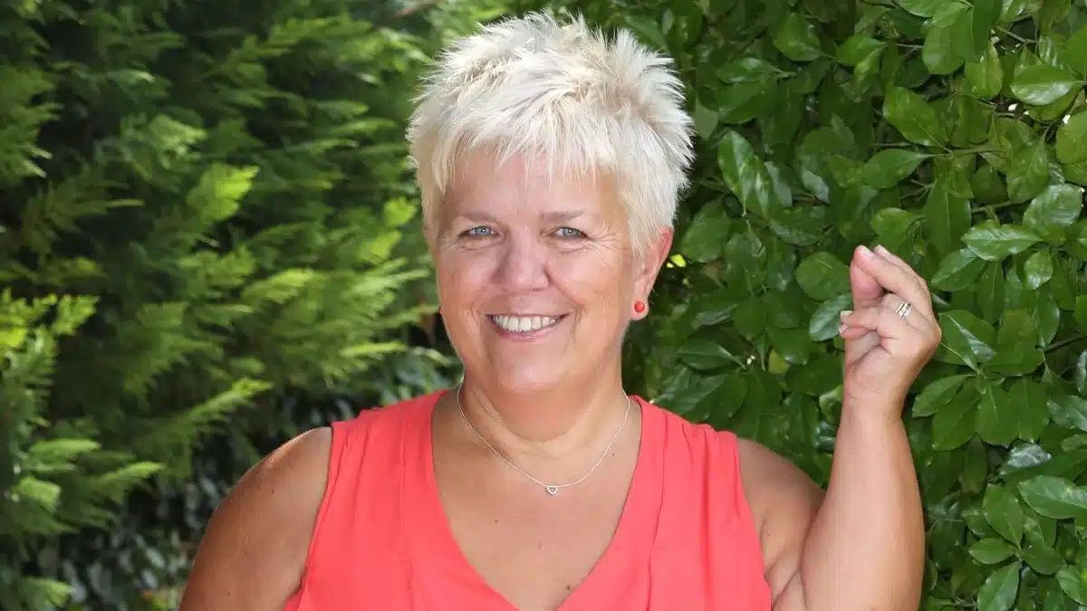 Mimie Mathy souriante devant un fond de verdure, cheveux courts blonds et haut corail