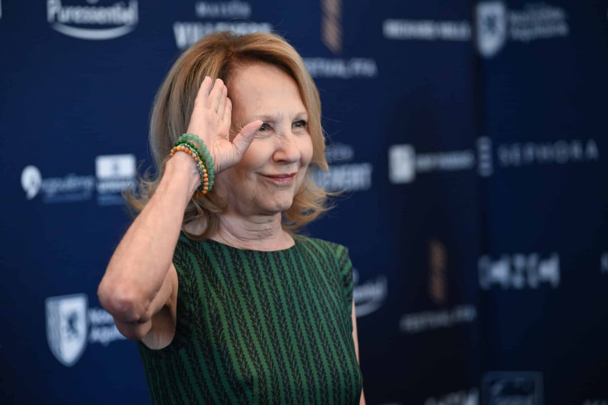 Nathalie Baye souriante lors d'un événement, levant la main devant un photocall bleu