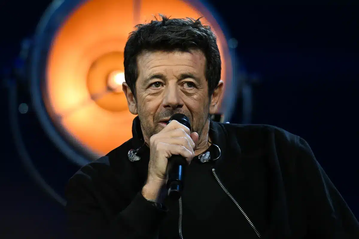 Patrick Bruel tenant un microphone sur scène, visage en gros plan sous un éclairage orangé