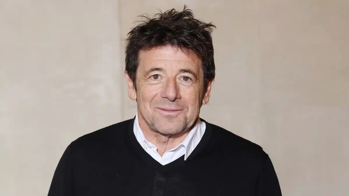 Patrick Bruel en portrait, pull noir col V sur fond beige, sourire discret
