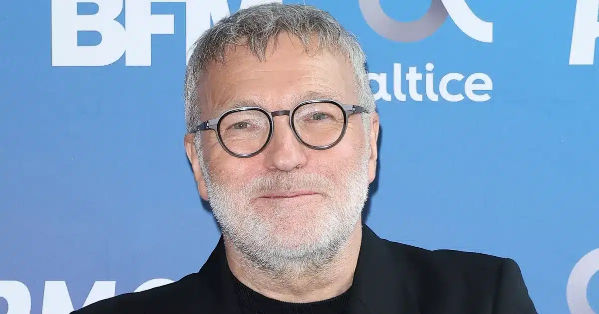 Laurent Ruquier en portrait, cheveux gris et lunettes rondes, devant un fond bleu BFM Altice