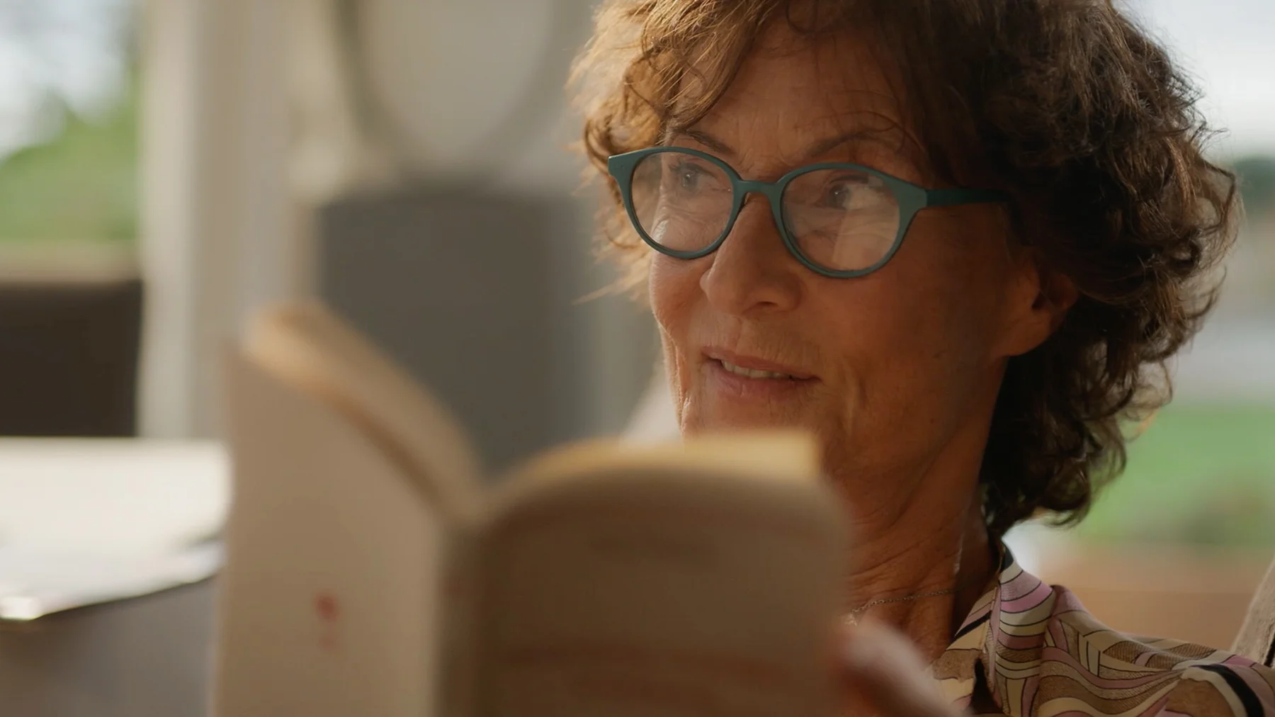 Une femme aux lunettes turquoise lit attentivement, ambiance chaleureuse et concentrée