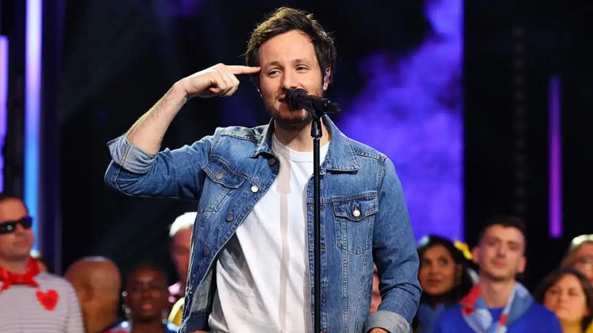 Vianney sur scène devant un micro, dans une veste en jean, pointant son doigt vers sa tempe